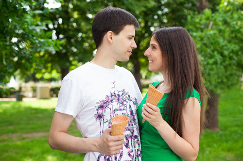 Chłopak z dziewczyną stoją w parku, patrzą na siebie i jedzą lody